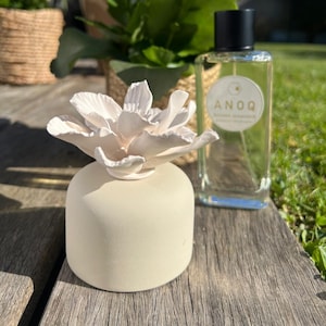 Puede incluir: Un difusor de cerámica color crema con una parte superior en forma de flor, junto a una botella de vidrio transparente con una tapa negra. La botella tiene una etiqueta blanca con la palabra "ANOQ". Los artículos están sobre una superficie de madera.