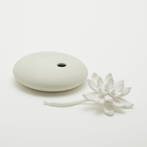 Op de afbeelding: Witte keramische diffuser met een ronde basis en een lotusbloem ontwerp. De diffuser heeft een klein gaatje in het midden van de basis en een wit koord bevestigd aan de bloem.