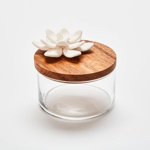 Op de afbeelding: Een glazen pot met een houten deksel en een witte keramische bloem erop. De pot is perfect om kleine spullen zoals sieraden of snuisterijen in op te bergen.