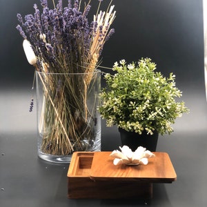 May include: A wooden box with a white ceramic flower on the lid. The box is open and has a clear glass vase inside with dried lavender and wheat stalks.