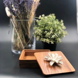 May include: A wooden box with a lid featuring a white ceramic flower design. The box is open and the lid is resting on top of the box. The box is made of dark wood with a natural grain pattern.