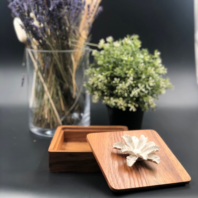 May include: A wooden box with a lid featuring a white ceramic flower design. The box is open and the lid is resting on top of the box. The box is made of dark wood with a natural grain pattern.