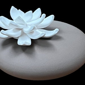 May include: A white ceramic flower sits atop a grey ceramic oval-shaped base. The flower has a delicate, intricate design.