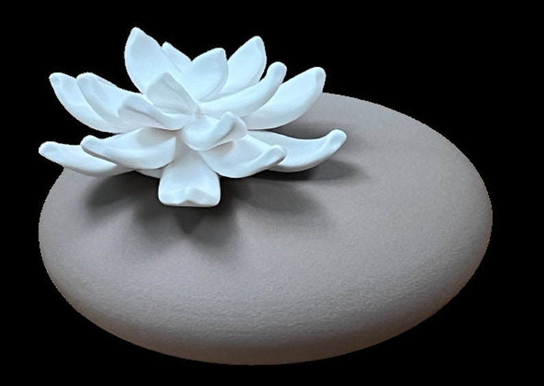 May include: A white ceramic flower sits atop a grey ceramic oval-shaped base. The flower has a delicate, intricate design.