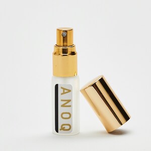 Può includere: Un piccolo flacone di vetro trasparente con un tappo dorato e un'etichetta dorata che dice "ANOQ". Il flacone è su una superficie bianca.