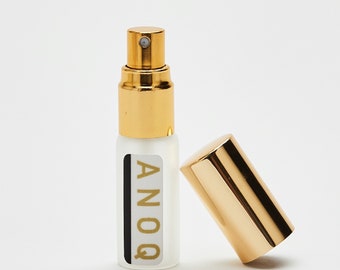 May include: A small, clear glass bottle with a gold cap and a gold label that says "ANOQ". The bottle is sitting on a white surface.