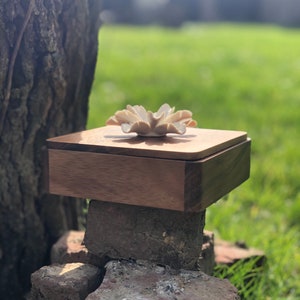 May include: A wooden box with a lid, featuring a white ceramic flower on top. The box is sitting on a stack of rocks in a grassy area.