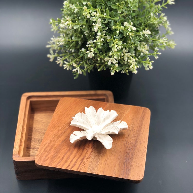 May include: A wooden box with a hinged lid. The lid has a white ceramic flower design on top. The box is open and the inside is empty.