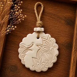May include: A circular off-white ceramic ornament featuring an intricate embossed floral and paisley pattern. The ornament has a scalloped edge and is suspended by a thick, natural-coloured jute twine loop knotted at the top for hanging.
