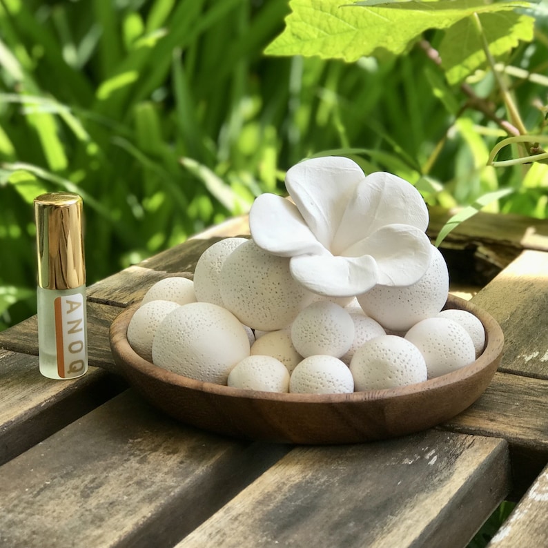 May include: White ceramic diffuser balls in a wooden bowl with a white ceramic flower on top. The diffuser balls are textured and the flower has five petals. A small bottle of essential oil is in the background.