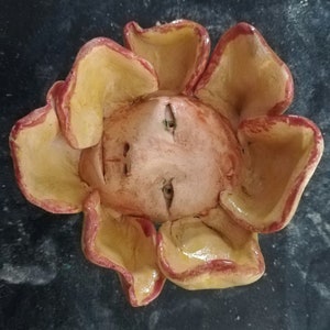 May include: A ceramic flower sculpture with a face in the centre. The flower is yellow with red edges and the face is a light orange colour.