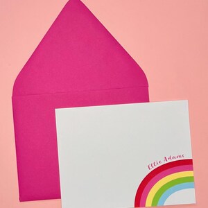 Op de afbeelding: Een witte kaart met een regenboogontwerp en de naam "Ellie Adams" in roze gedrukt. De kaart wordt geleverd met een roze envelop.
