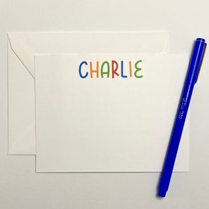 May include: A white card with the name "CHARLIE" written in colorful letters. The letters are in different colors: blue, orange, red, green, and light blue. A blue pen is lying next to the card.