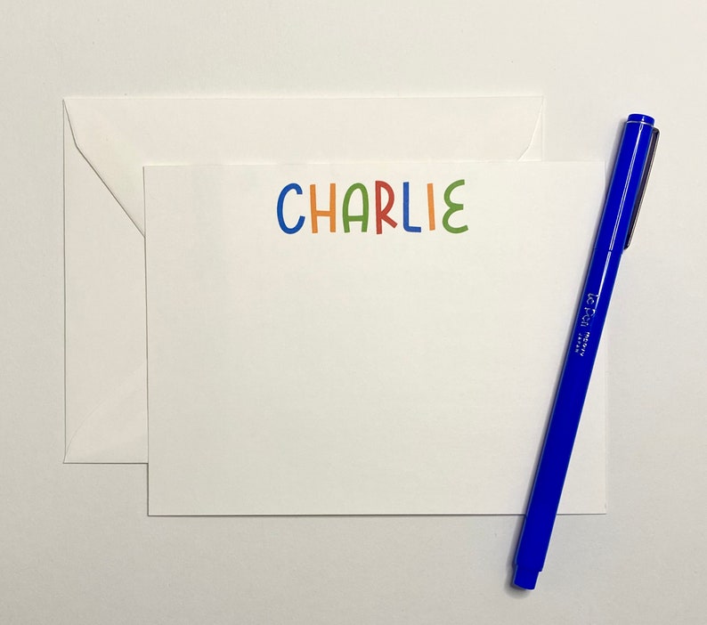 May include: A white card with the name "CHARLIE" written in colorful letters. The letters are in different colors: blue, orange, red, green, and light blue. A blue pen is lying next to the card.