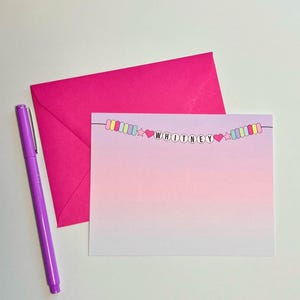 May include: A pink and purple ombre card with a colorful beaded garland that spells out the name "WHITNEY".