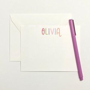 May include: A white card with a pink envelope and a purple pen. The card has the name "Olivia" written on it in colorful letters.