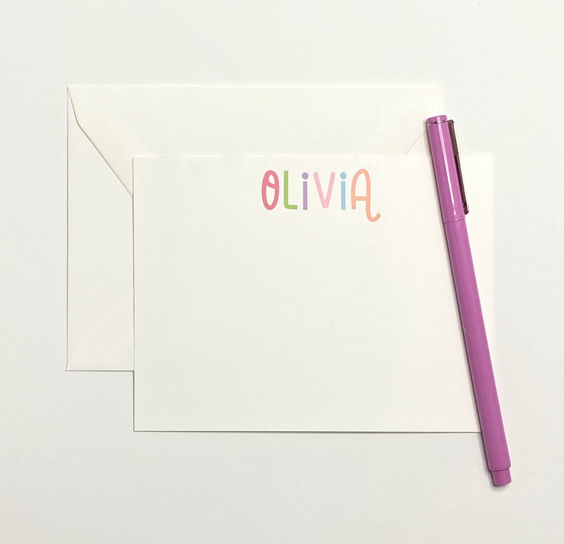 May include: A white card with a pink envelope and a purple pen. The card has the name "Olivia" written on it in colorful letters.