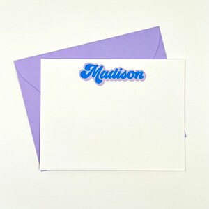 May include: A white card with a purple envelope. The card has the name "Madison" written in blue with a retro font.