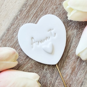May include: White ceramic heart-shaped cake topper with the word "Aunt" written in cursive and a small heart in the center.
