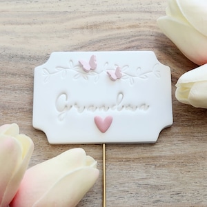 May include: White cake topper with the word "Grandma" in cursive, two pink butterflies, and a pink heart. The topper is on a gold stick.