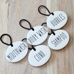 May include: Five white oval ceramic bathroom labels with black string hangers. The labels read "Mouthwash", "Body Wash", "Cond.", "Shampoo", and "Bubbles".