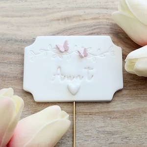 May include: A white rectangular sign with the word "Aunt" in cursive, adorned with two pink butterflies and a heart. The sign is attached to a gold-colored metal stake, perfect for a floral arrangement or gift.