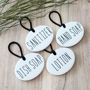 May include: Four white oval ceramic labels with black elastic loops and black text. The labels read "Sanitizer", "Hand Soap", "Dish Soap", and "Lotion".