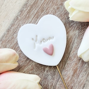 May include: White ceramic heart with the word "Mom" embossed on it. A small pink heart is centered below the word. The heart is attached to a gold-colored stick.