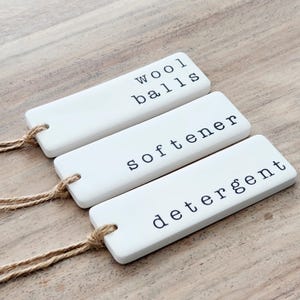 May include: Three white ceramic labels with twine attached. The labels are for laundry items and read "wool balls", "softener", and "detergent".