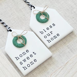 May include: Two white wooden house-shaped signs with green wreaths and twine bows. The signs have black text that reads "home sweet home" and "bless our home".