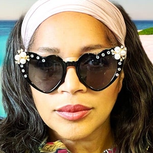 Könnte beinhalten: Eine Frau mit einem rosafarbenen Stirnband und schwarzen herzförmigen Sonnenbrillen mit Perlenbesatz. Sie trägt eine Bluse mit Blumenmuster.