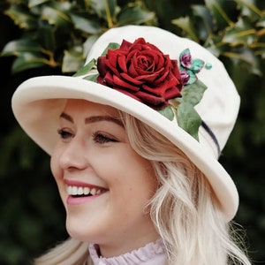 May include: A white felt hat with a wide brim and a red rose embellishment. The hat has a green ribbon around the crown.