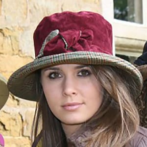 May include: A burgundy velvet hat with a plaid trim and a decorative bow. The hat has a wide brim and is worn by a person with long brown hair.