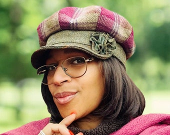 May include: A woman wearing a green and pink plaid newsboy cap with a brown flower accent. She is wearing a pink shawl and has red nail polish.