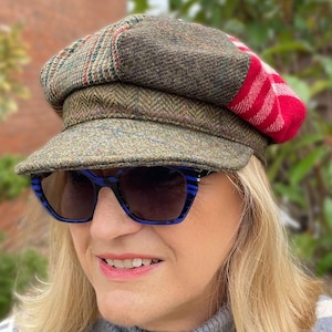 May include: A tweed newsboy cap with a red and white plaid accent. The cap is olive green with a herringbone pattern. The person is wearing blue and black striped sunglasses.