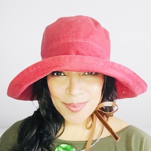 May include: A woman wearing a red wide-brimmed hat with a brown velvet ribbon tied around the crown. She is wearing a green shirt and a large green gemstone necklace.