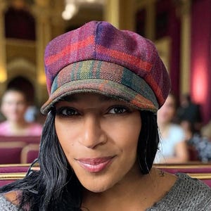 Könnte beinhalten: Eine mehrfarbige karierte Newsboy-Mütze mit runder Krone und kurzem Schirm. Die Mütze hat eine Mischung aus lila, roten, grünen und braunen Farbtönen. Der Hut ist aus einem strukturierten Stoff gefertigt.