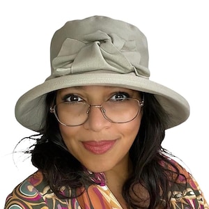 Puede incluir: Una mujer lleva un sombrero de sol gris claro con un gran lazo en la parte superior. El sombrero tiene un ala ancha y está hecho de una tela suave y texturizada. También lleva gafas redondas.