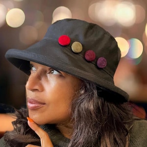 May include: A black bucket hat with a wide brim and a row of colourful felt pom-poms on the side. The pom-poms are red, green, burgundy, and purple.