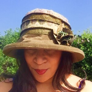 Puede incluir: Un sombrero de ala ancha de color verde, marr&oacute;n y blanco con un adorno floral. El sombrero est&aacute; hecho de tejido tejido y tiene una corona ligeramente ca&iacute;da.