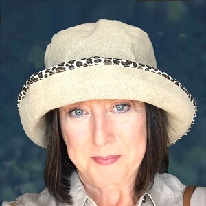 May include: A woman wearing a beige linen hat with a brown and black leopard print band. The hat has a wide brim and is slightly tilted on her head. She is wearing a light beige shirt.