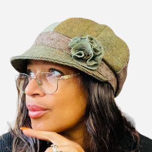 May include: A green and brown tweed newsboy hat with a decorative flower. The hat has a small brim and a mix of textures and patterns, offering a vintage aesthetic. The hat is a stylish accessory.