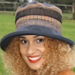 Può includere: Una donna con i capelli biondi ricci indossa un cappello con motivo blu e marrone. Sorride e guarda la telecamera. Indossa una giacca marrone.