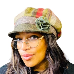May include: A woman wearing a green, red, and brown plaid newsboy cap with a green fabric flower accent. The cap has a wide brim and is made of a soft, textured fabric.