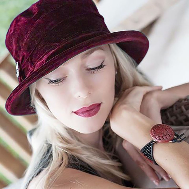 May include: A woman wearing a burgundy velvet cloche hat with a small decorative button on the side. She is wearing a black bracelet with a red, carved, circular charm.