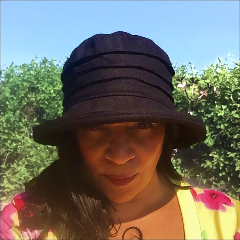Puede incluir: Un sombrero de sol negro de ala ancha con una corona plisada. El sombrero lo lleva una persona con el pelo oscuro y una blusa de estampado floral.