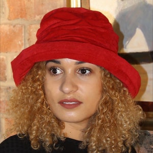 May include: A woman with curly blonde hair is wearing a red suede hat. The hat has a wide brim and a slightly slouched crown.
