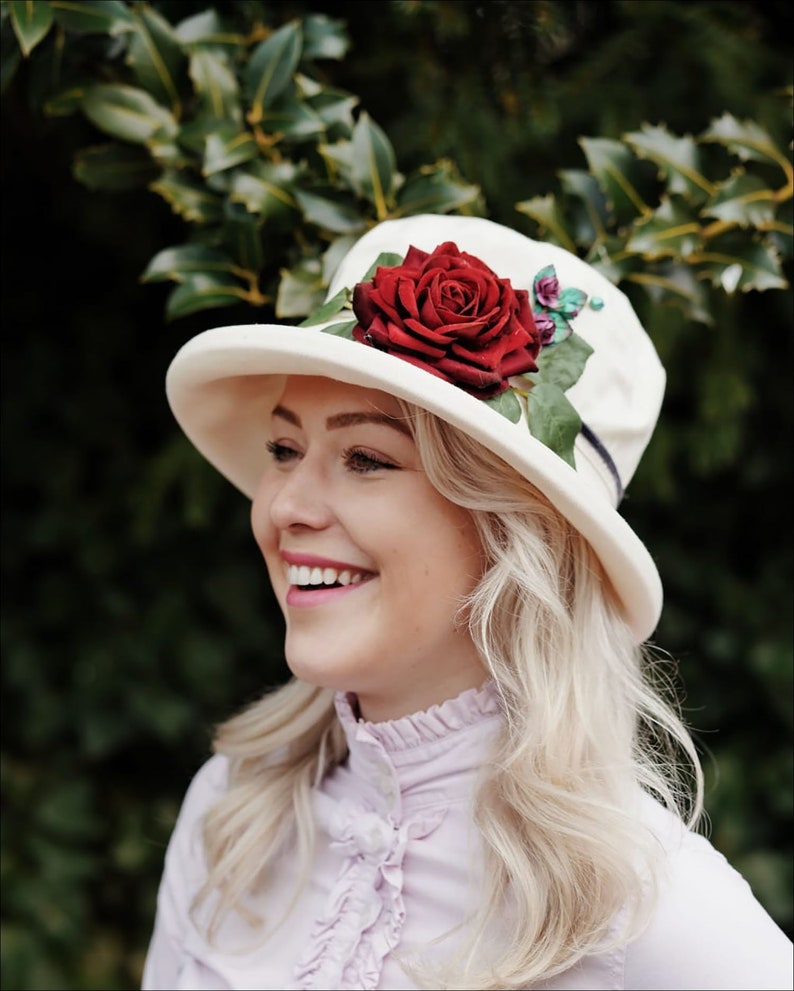 May include: A white felt hat with a wide brim and a red rose embellishment. The hat has a green ribbon around the crown.