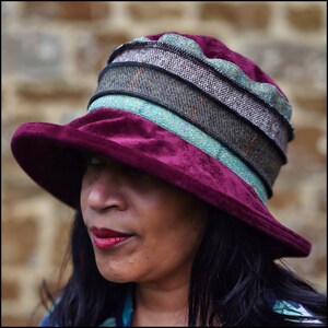 Puede incluir: Un sombrero de terciopelo burdeos con una ala ancha y una banda estampada multicolor. El sombrero est&aacute; hecho de una tela suave y lujosa y tiene un aspecto elegante y vintage.