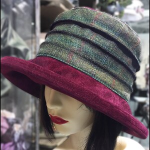 Puede incluir: Un sombrero de fieltro burdeos con una banda de cuadros verdes y negros. El sombrero tiene un ala ancha y una corona suave y flexible.